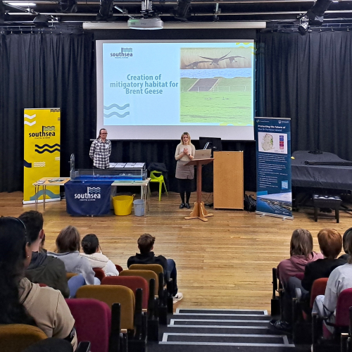 Women and man stood in an auditorium in front of a group of students and large screen with a presentation on it 