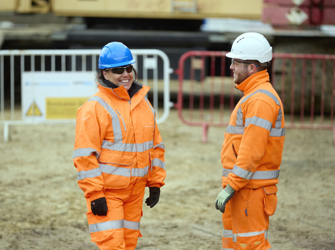 man and women on a construction site wearing orange hi vis work wear, both smiling with the man waving his arms in the air