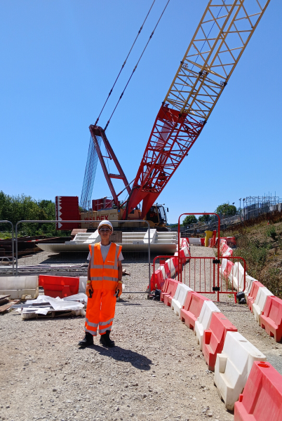 Young man stood in front of a crane wearing orange hi-vis workwear