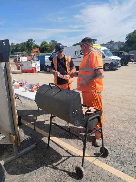 Construction workers in orange hi-vis workwear stood cooking at a BBQ