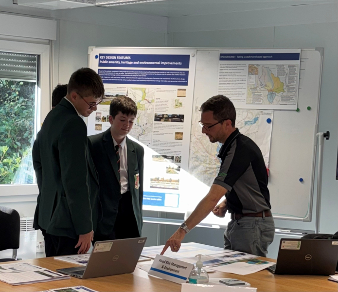 Two school boys talking to a male in a site cabin stood by a table with a laptop and project information