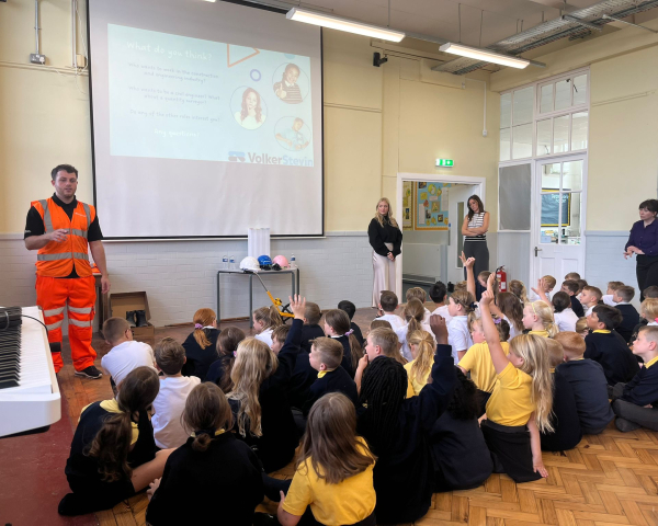 Man in hi-vis orange workwear stood in classroom presenting to children sat down on floor