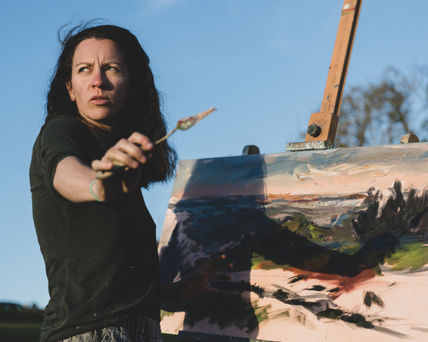 Women artist holding a paint brush stood in front of a picture on an easel
