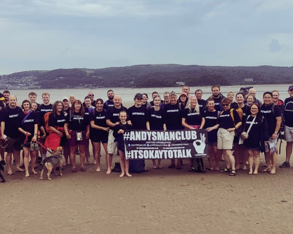 group of men and women stood together on a beach holding a black banner that says Andy's Man Club