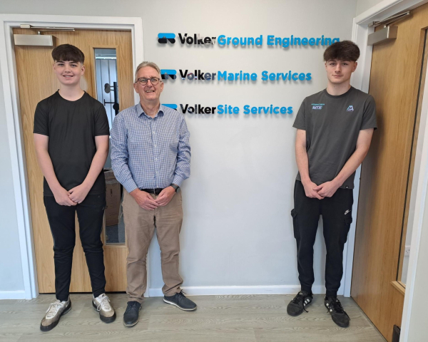 Three men stood in front of a white wall with three logos on the wall behind them