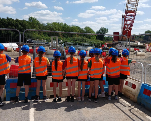 Group of school children in orange hi-vis wear and blue hard hats stood by fencing on a construction site