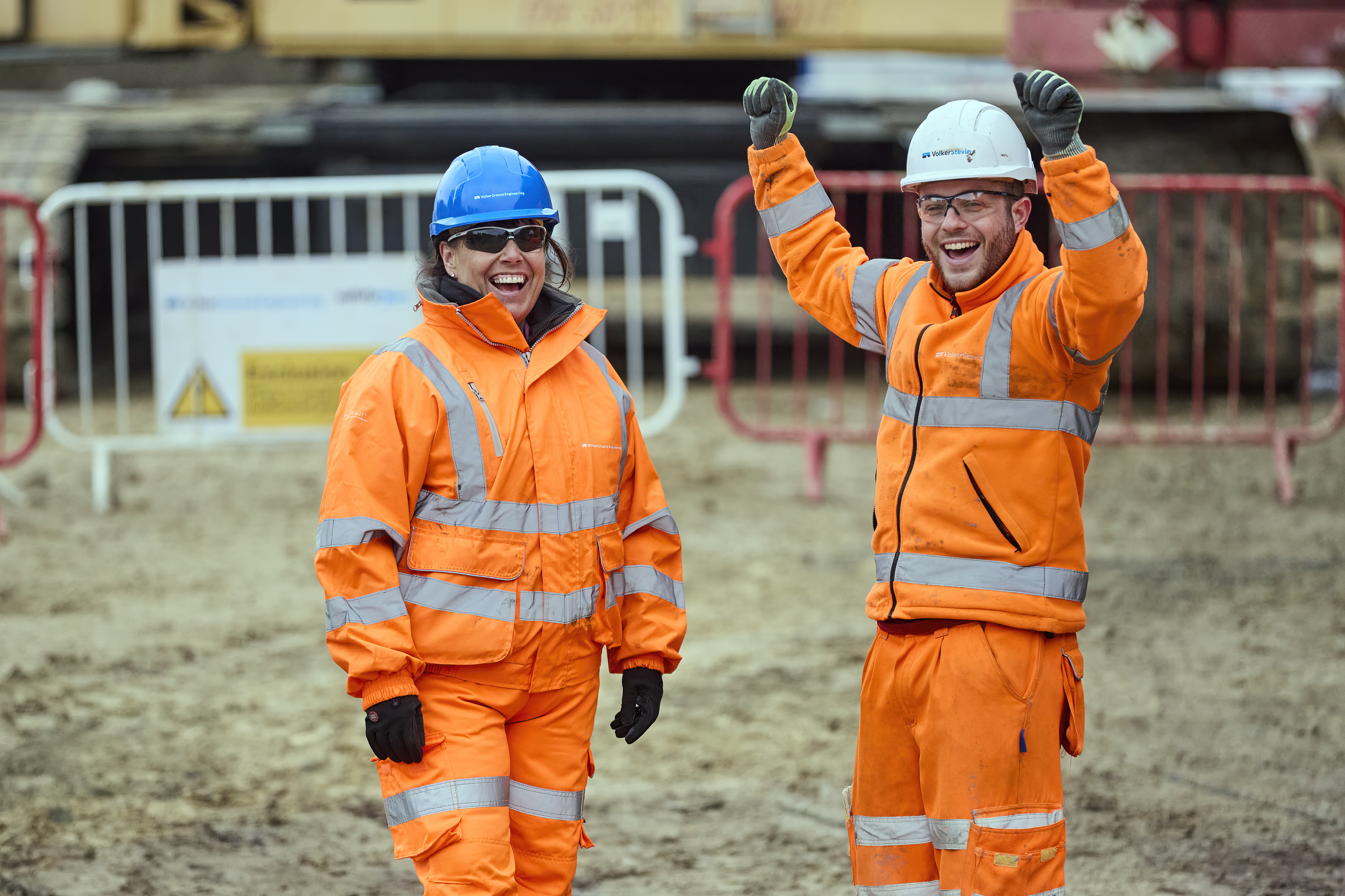 man and women on a construction site wearing orange hi vis work wear, both smiling with the man waving his arms in the air