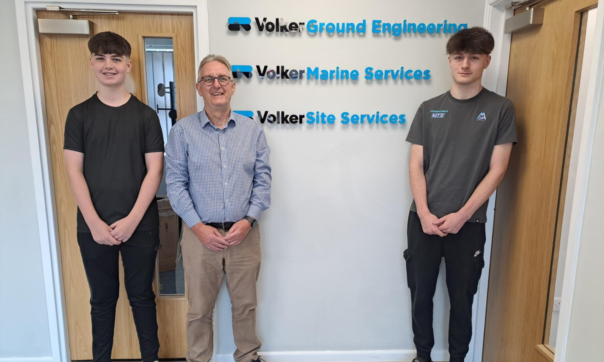 Three men stood in front of a white wall with three logos on the wall behind them