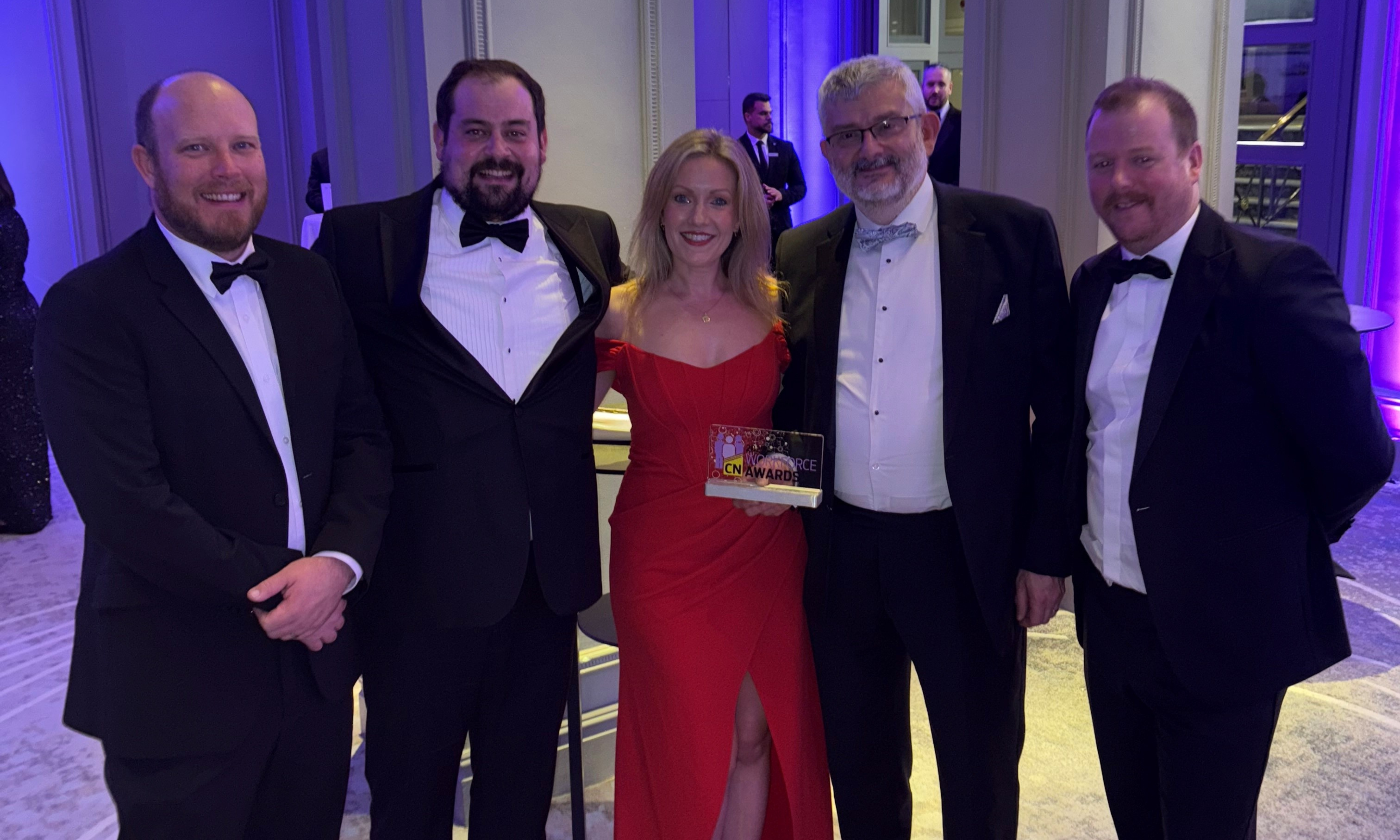 Three men in black tie and one women in a red dress standing with a glass trophy